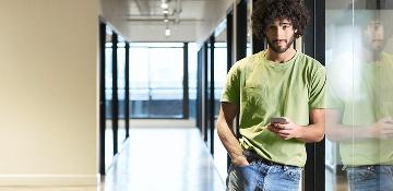 Ein junger Mann steht angelehnt im Gang eines Bürogebäudes und schaut von seinem Smartphone auf.