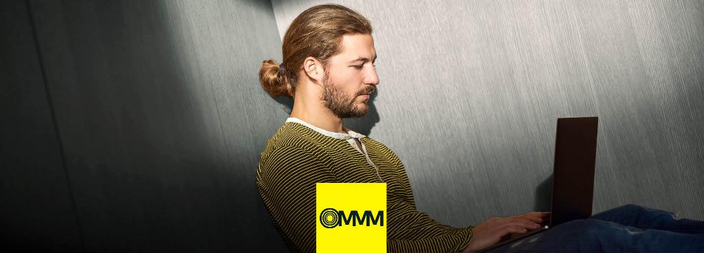 Ein junger, bärtiger Mann mit Pferdeschwanz sitzt auf dem Boden mit einem Laptop auf seinen Knien.
