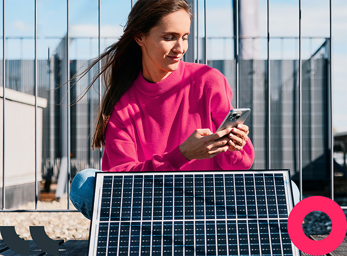 Eine Frau sitzt neben einem Solarpanel auf dem Dach und blickt l&auml;chelnd auf ihr Smartphone.

 