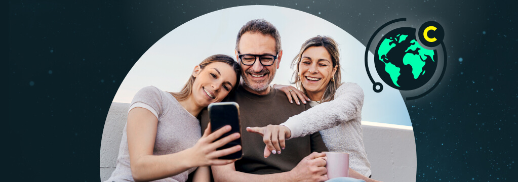 Vater, Mutter und Tochter sitzen l&auml;chelnd zusammen und schauen auf ein Handy, das die Tochter h&auml;lt.
 