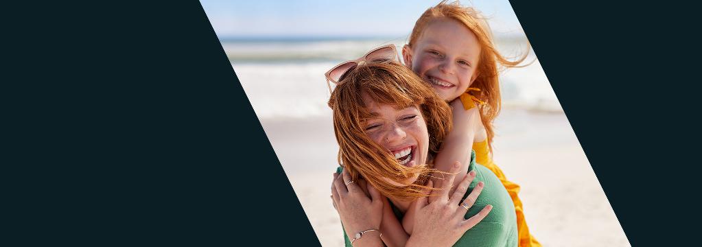 Eine lachende Frau tr&auml;gt ein Kind huckepack am Strand. Im Hintergrund sieht man das Meer.