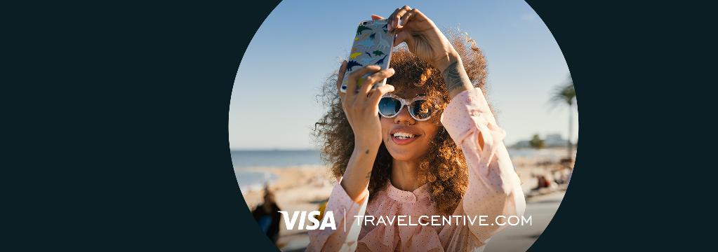 Eine junge Frau mit Sonnenbrille steht lachend am Strand und macht ein Selfie mit ihrem Smartphone.